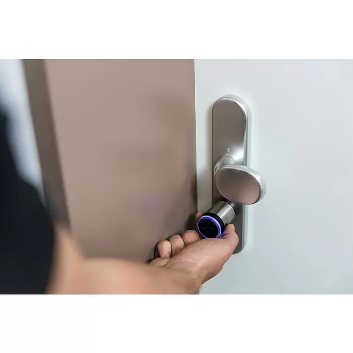 Abus Elektronisches Türschloss CYLOX™ One - Bedienung: Über App, Mit Schließung innen, Baulänge innen: 45 mm - 55 mm - Eine Hand bedient einen intelligenten Türschließzylinder aus Metall mit einem leuchtenden violetten Ring, der an einem silbernen Türgriff befestigt ist.