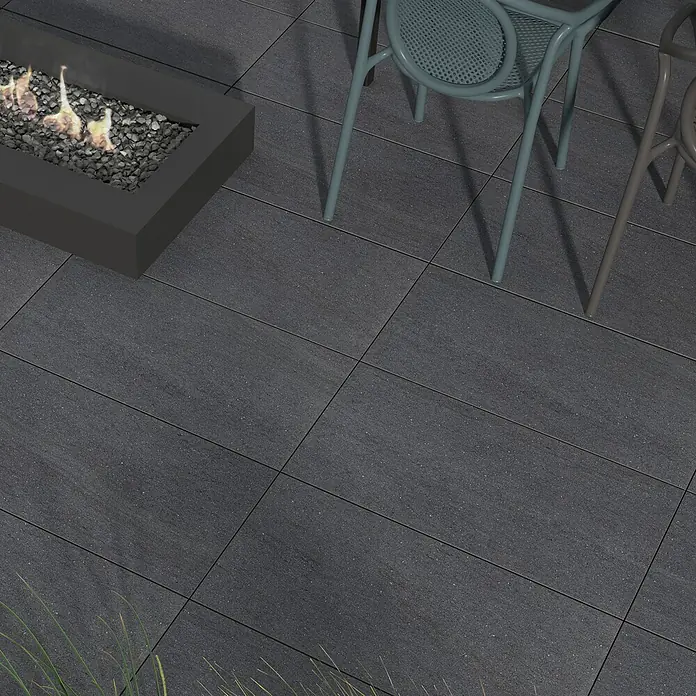 Terrassenfliese Kamari 2.0 - 39,7 x 79,7 x 2 cm, Anthrazit, Matt - Das Bild zeigt eine moderne Terrasse mit dunklen Pflastersteinen, einem stilvollen Feuerkorb und bequemen Gartenstühlen für gemütliche Abende im Freien.