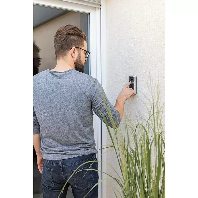 Abus HomeTec Pro Fingerabdruck Lesegerät CFS3100S - L x B x H: 29 x 67 x 143 mm, Schwarz / Silber - Mann drückt Taste an einem silber-schwarzen Smart-Türklingel/Zutrittssystem an einer Wand.