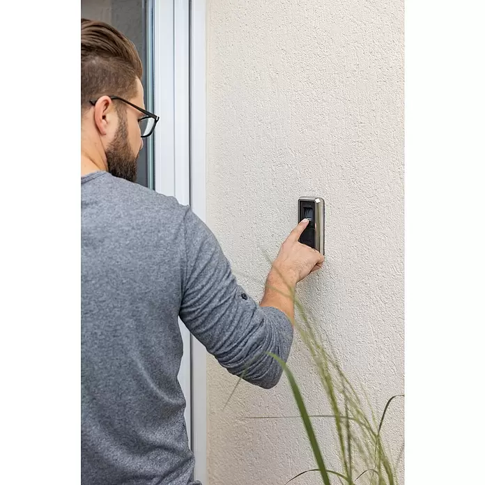 Abus HomeTec Pro Fingerabdruck Lesegerät CFS3100S - L x B x H: 29 x 67 x 143 mm, Schwarz / Silber - Ein Mann nutzt einen silber-schwarzen Fingerscanner an einer Wand neben einer Tür und legt seinen Finger zur Zugangskontrolle darauf.