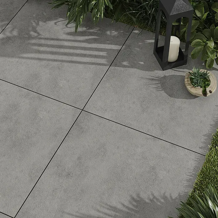 Cersanit Terrassenfliese Metropolis - 59,3 x 59,3 x 2 cm, Grey, Matt - Graue Terrassenplatten aus Betonstein mit schmalen Fugen, umgeben von Pflanzen und einer Dekolaterne.