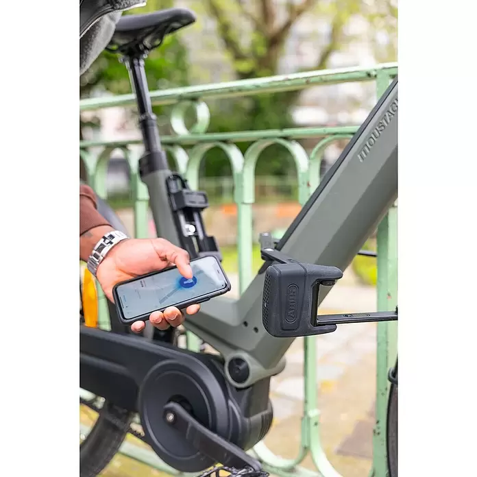 Abus Faltschloss BORDO™ One 6000A/120 - Länge: 120 cm - Eine Person steuert eine schwarze Abus Smart Lock, die in ein olivfarbenes Moustache E-Bike integriert ist, per Smartphone-App.