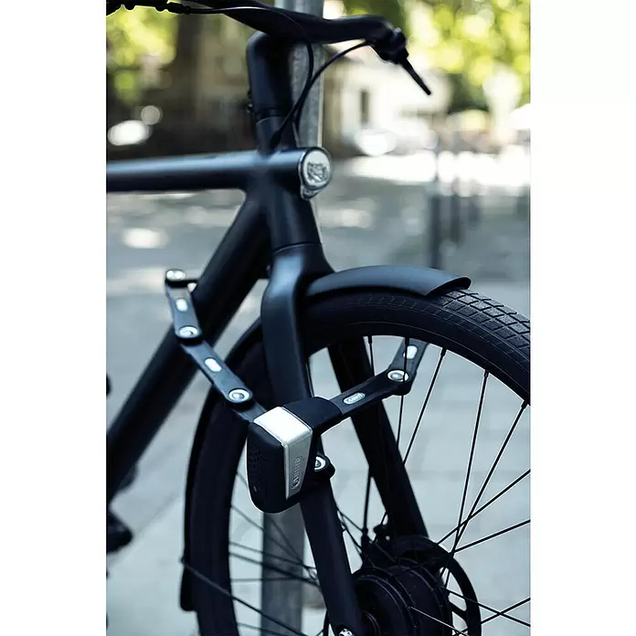 Abus Faltschloss BORDO™ One 6500A/110 - Länge: 110 cm - Ein schwarzes Fahrrad mit einem schwarz-silbernen Faltschloss an Vorderrad und Rahmen, an einem Metallpfosten auf einer Straße gesichert.