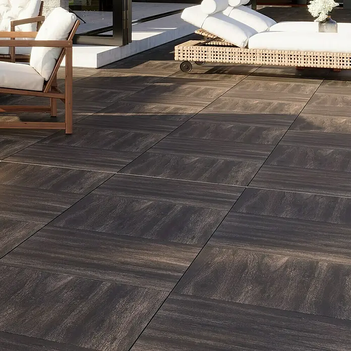 Terrassenfliese Strobus - 60 x 60 x 2 cm, Ebony/Braun, Matt - Nahaufnahme von dunklen Bodenfliesen auf einer Terrasse mit Loungemöbeln im Hintergrund. Die Fliesen haben eine natürliche Holzoptik.
