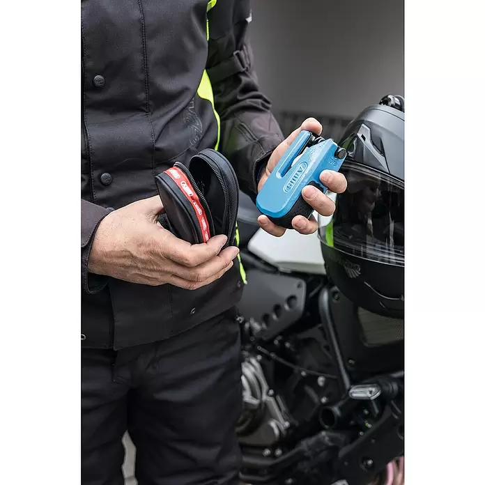 Abus Bremsscheibenschloss GRANIT™ Detecto One 8078 2.0 - Geeignet für: Motorrad, Blau - Eine Person in Motorradausrüstung hält ein blaues ABUS Bremsscheibenschloss und seine schwarze Tragetasche, neben Motorradhelm und Bike.