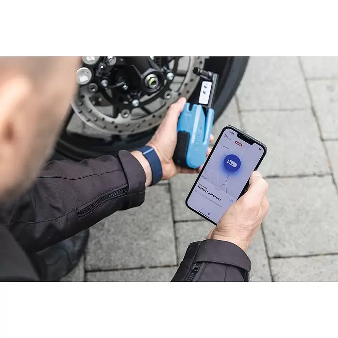 Abus Bremsscheibenschloss GRANIT™ Detecto One 8078 2.0 - Geeignet für: Motorrad, Blau - Eine Person hält ein blaues Motorrad-Bremsscheibenschloss und ein Smartphone mit dessen Standort auf einer Karte, nahe einem Motorradrad.