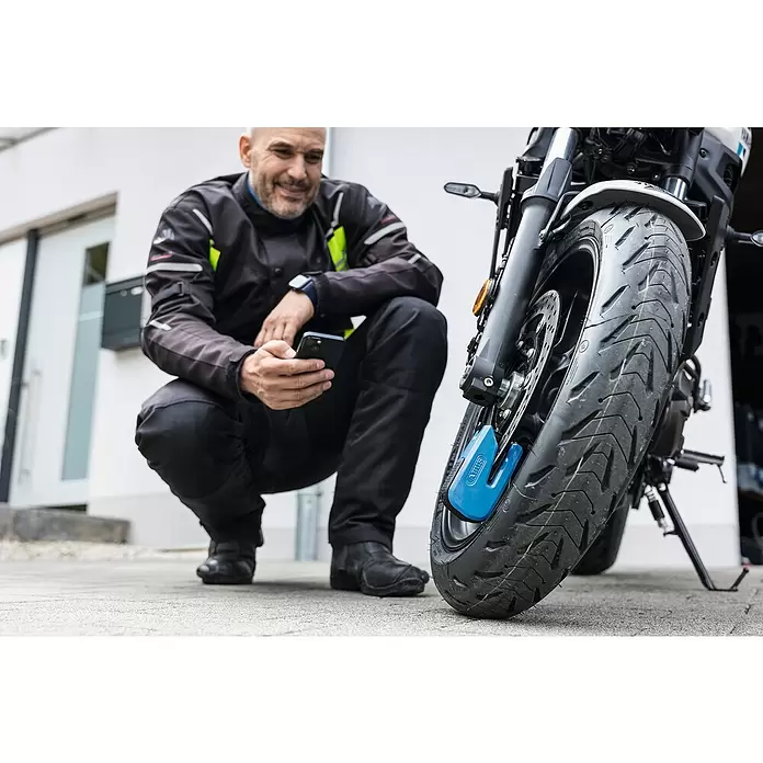 Abus Bremsscheibenschloss GRANIT™ Detecto One 8078 2.0 - Geeignet für: Motorrad, Blau - Ein Mann hockt neben einem Motorrad mit einem blauen Bremsscheibenschloss am Vorderrad und bedient sein Telefon.
