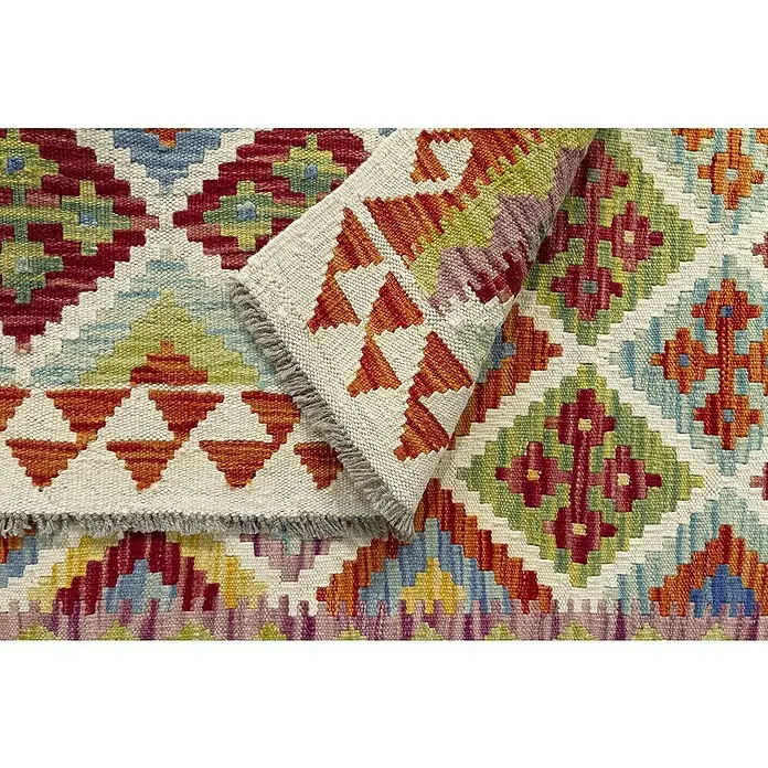 Alfombra de pelo corto Kilim Herat - Multicolor, 180 x 120 cm, 100% lana - Alfombra colorida con patrones geométricos. La alfombra tiene flecos y está parcialmente doblada, revelando más patrones.