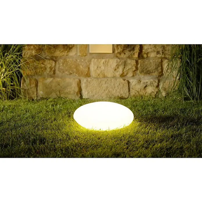 LED-Solarstein - Weiß, L x B x H: 30 x 40 x 16 cm - Eine runde, leuchtende Gartenleuchte in Form eines Steins auf Rasen vor einer Steinmauer. Das Licht erzeugt eine angenehme Atmosphäre im Garten.