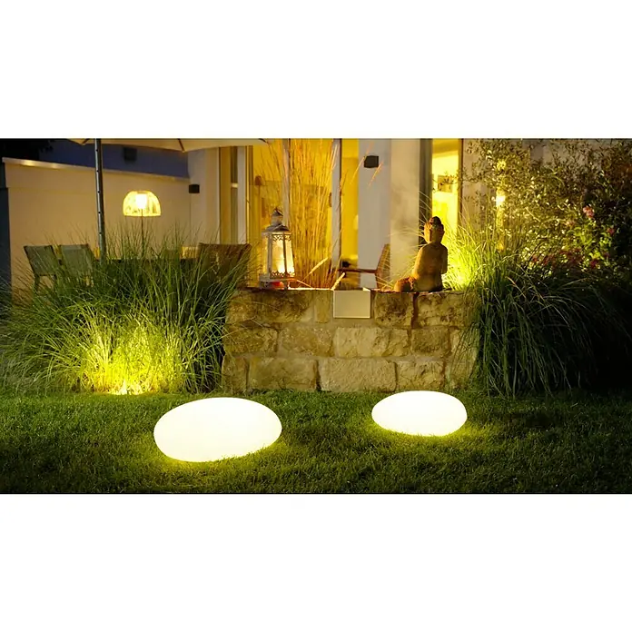 Solar-Dekoleuchte - Weiß, L x B x H: 36 x 50 x 19,5 cm - Garten mit zwei leuchtenden Steinen, Buddha Statue und Laterne auf einer Steinmauer, umgeben von Gräsern und Pflanzen.