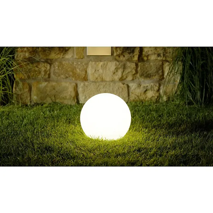 LED-Solar-DekoleuchteWeiß, 30 x 27 cm - Eine Gartenleuchte in Kugelform strahlt helles Licht auf den Rasen, während eine Steinmauer im Hintergrund für eine stimmungsvolle Atmosphäre sorgt.