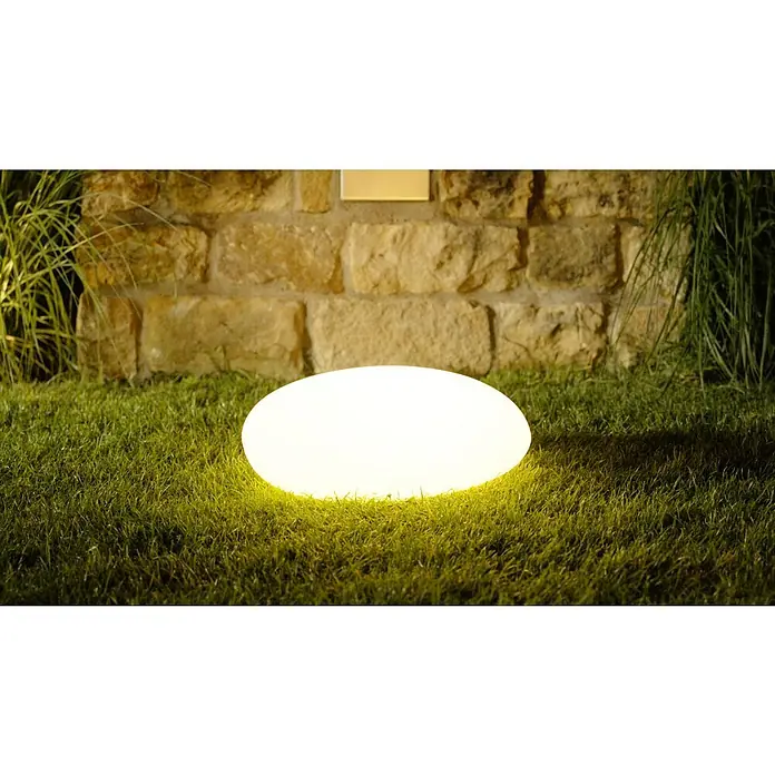 Solar-Dekoleuchte - Weiß, L x B x H: 36 x 50 x 19,5 cm - Eine leuchtende, ovale Gartenleuchte sitzt auf einem grünen Rasen vor einer steinernen Wand und sorgt für eine stimmungsvolle Atmosphäre.