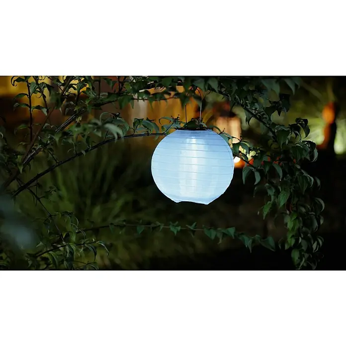 BAUHAUS Solar-Hängeleuchte Lampion - 0,06 W, Weiß, IP44, Durchmesser: 20 cm - Eine leuchtende Papierlaterne hängt an einem blühenden Baum und erhellt sanft die Umgebung mit einem blauen Lichtschein.