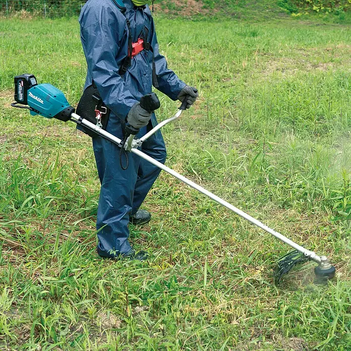 Makita XGT 40V max. Akku-Freischneider UR006GM102 - 40 V, 4 Ah, 1 Akku, Schnittbreite Faden: 43 cm, Schnittbreite Messer: 25,5 cm - Person, die einen Makita Akku-Rasentrimmer in einem Feld mit hohem Gras verwendet, mit blauem Anzug, der für den Job geeignet ist.