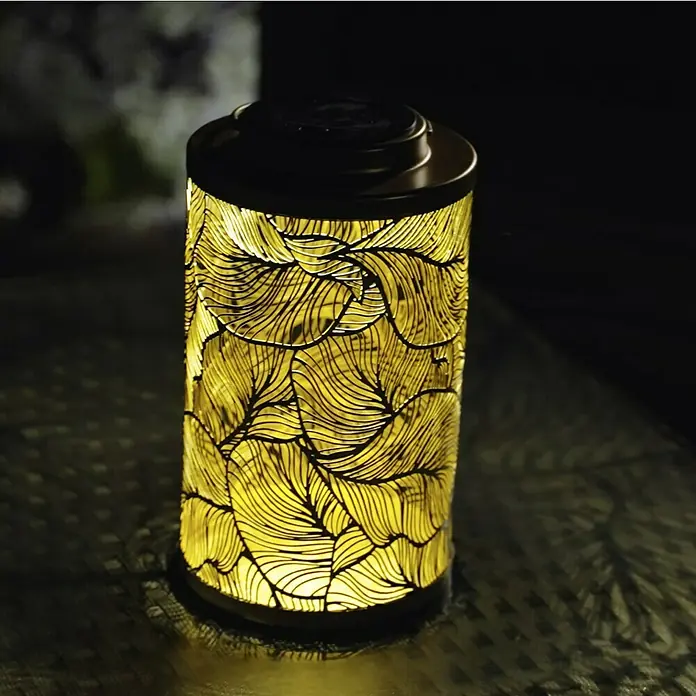 BAUHAUS Solar-Tischleuchte Dekolaterne - Gold - Nahaufnahme einer zylindrischen Solarleuchte mit Blattmuster, die ein warmes, gelbes Licht abstrahlt und eine gemütliche Atmosphäre schafft.