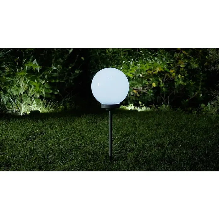 BAUHAUS Solar-Dekoleuchte Fara - Durchmesser: 25 cm, Höhe: 67,7 cm, LED - Nachts beleuchtet eine kugelförmige Gartenleuchte mit Stab einen Rasen, umgeben von Sträuchern. Sie sorgt für stimmungsvolle Gartenbeleuchtung.