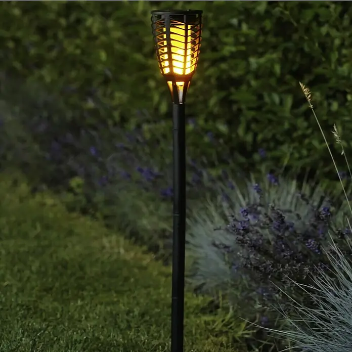 BAUHAUS Solar-Erdspieß Fackel Flame - Schwarz - Eine Solar-Gartenfackel mit flackerndem Licht steht inmitten von grüner Vegetation und blühenden Pflanzen.