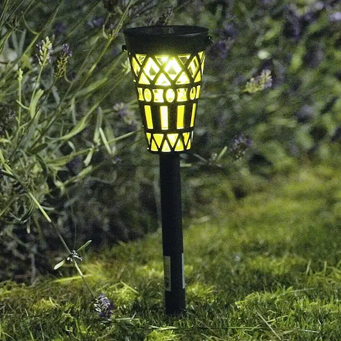 BAUHAUS Solar-Erdspieß - Schwarz/Gold, IP44, 3 lm - Nahaufnahme einer Solar-Gartenfackel mit dekorativem Design, die im Gras vor Büschen mit lila Blüten steht.