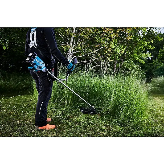 Makita LXT 18V Akku-Freischneider DUR369AZX6 - 18 V, Ohne Akku, Schnittbreite Faden: 43 cm, Schnittbreite Messer: 25,5 cm - Eine Person trimmt Gras mit einem Makita Akku-Rasentrimmer mit Tragegurt vor Bäumen und Büschen.
