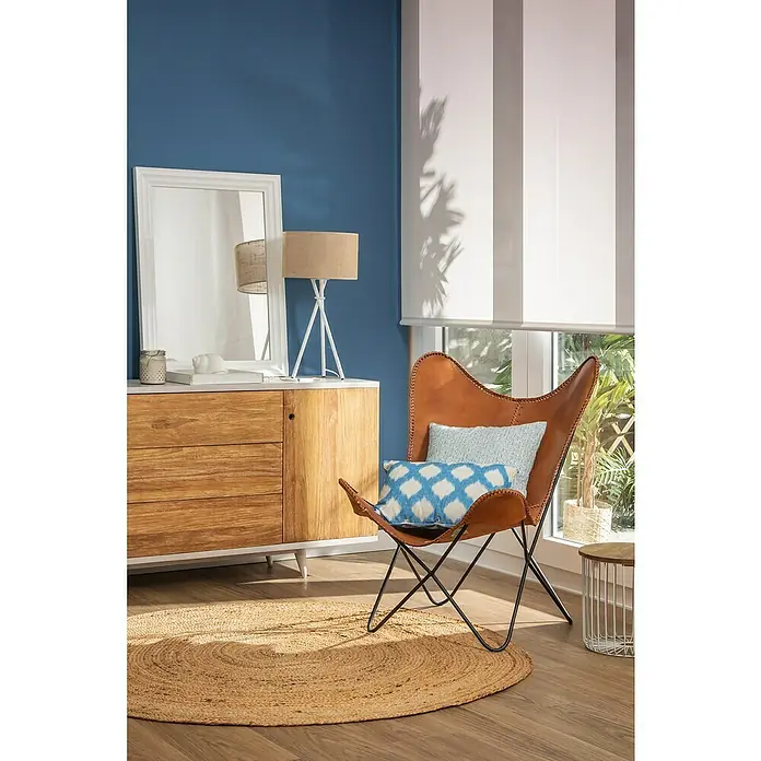 Alfombra de yute Nazar - Beige, Diámetro: 100 cm - Interior con sillón mariposa de cuero marrón, cojines azules sobre una alfombra redonda, cómoda con espejo y estores enrollables.