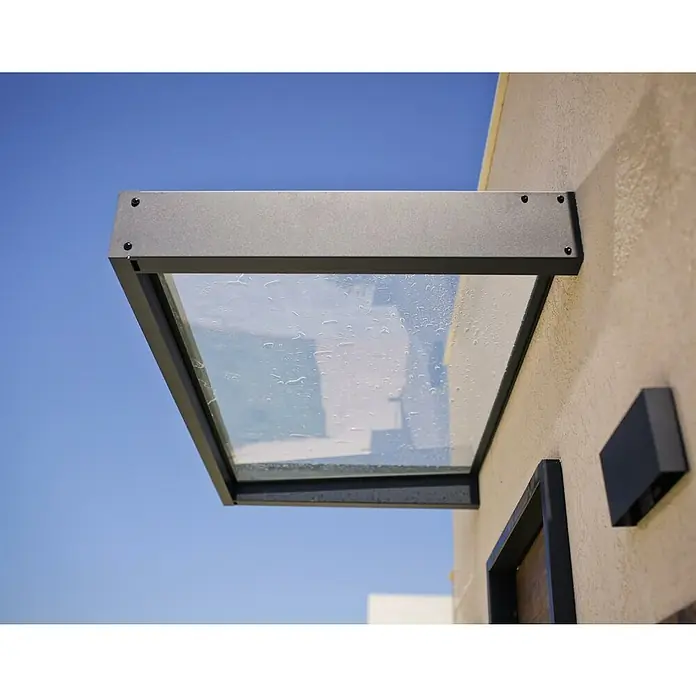 Palram – Canopia Marquesina para puerta Sophia - 160 x 95 x 17 cm, Gris antracita, Color del techo: Blanco opal - Primer plano de una marquesina de vidrio gris moderna fijada al exterior de un edificio, con gotas de lluvia en la superficie del vidrio.