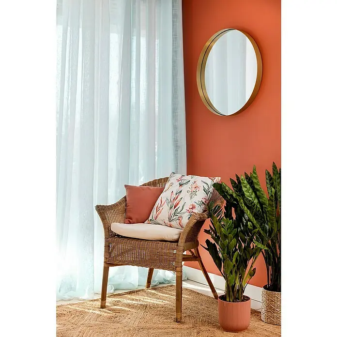 Espejo de pared Gia con marco - Diámetro: 60 cm, Oro - Sillón decorativo de ratán con cojines y plantas junto a un espejo. La disposición crea un ambiente acogedor y elegante.