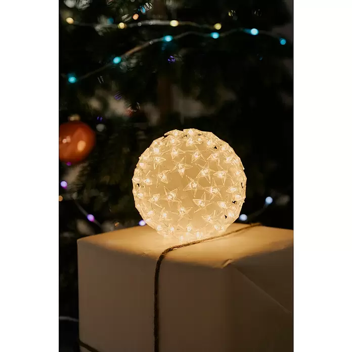 Tween Light LED-Weihnachtsdekoleuchte Sterne - Weiß, Netzbetrieben, LED, 13 cm - Leuchtende Kugel aus klaren Sternen auf einem weißen Geschenkpaket, vor verschwommenem Weihnachtsbaum.
