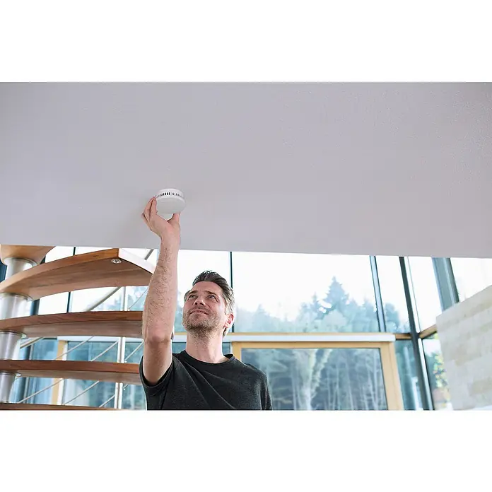 Abus Rauchwarnmelder-Set RWM90 - Alarmsignal: 85 dB, 3 Stk., Höhe: 36 mm - Mann installiert einen Rauchmelder an der Decke, um die Sicherheit im Haus zu gewährleisten. Der Rauchmelder ist weiß und rund.