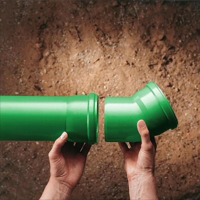 KG Bogen KG 2000 - Nennweite: 110 mm, 45 ° - Bild einer grünen Kunststoff-Abwasserrohrverbindung, die von zwei Händen vor einem Hintergrund aus Erde gehalten wird.