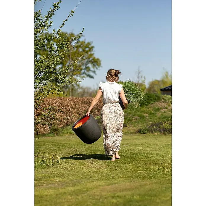 Capi Europe Urban Pflanztopf Smooth - Außenmaß (Ø x H): 34 x 35 cm, Schwarz, Kunststoff - Frau trägt einen stilvollen, runden Pflanztopf von bellavista in Schwarz und Orange durch den Garten.