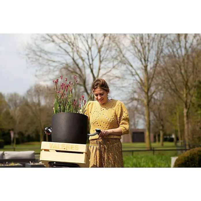 Capi Europe Urban Pflanztopf Smooth - Außenmaß (Ø x H): 34 x 35 cm, Schwarz, Kunststoff - Eine Frau betrachtet einen schwarzen Pflanztopf mit rosafarbenen Blumen auf einem Fahrrad, der in einer Holzkiste mit der Aufschrift "VALERES" steht.