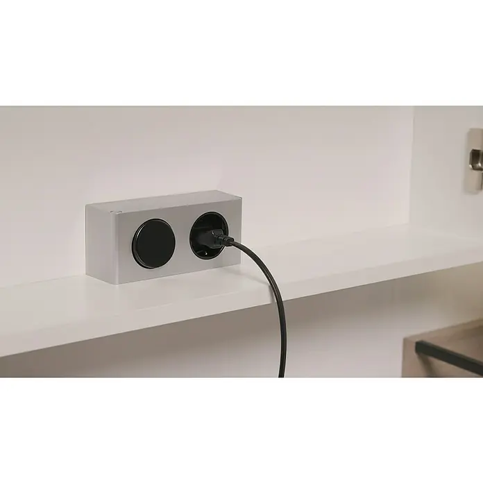 Riva LED-Spiegelschrank Luciana - B x H: 72 x 69,6 cm, Holz, Weiß - Eine moderne, silberfarbene Steckdosenleiste mit zwei Anschlüssen, von denen einer mit einem schwarzen Stecker belegt ist, auf einem weißen Regal.