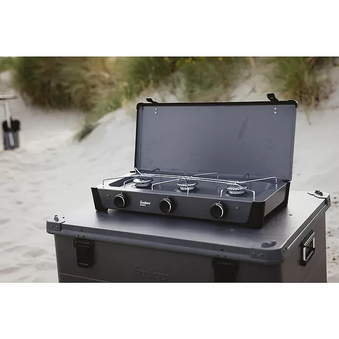 Enders Gaskocher Bondy 3Z - 61 x 32 x 33 cm, 2,3 kW, Stahl, Anzahl Brenner: 3 Stk., mit Zündsicherung - Dunkelgrauer Enders Campingkocher mit drei Brennern, geöffnet, auf passender Aufbewahrungsbox im sandigen Außenbereich.