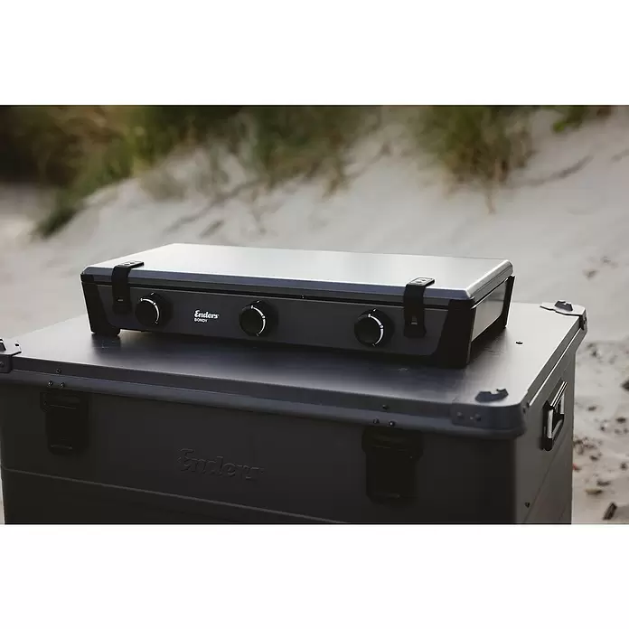 Enders Gaskocher Bondy 3Z - 61 x 32 x 33 cm, 2,3 kW, Stahl, Anzahl Brenner: 3 Stk., mit Zündsicherung - Dunkelgrauer Enders Campingkocher "Bondy" mit drei Brennern, auf passender Enders Aufbewahrungsbox im Freien auf Sand stehend.
