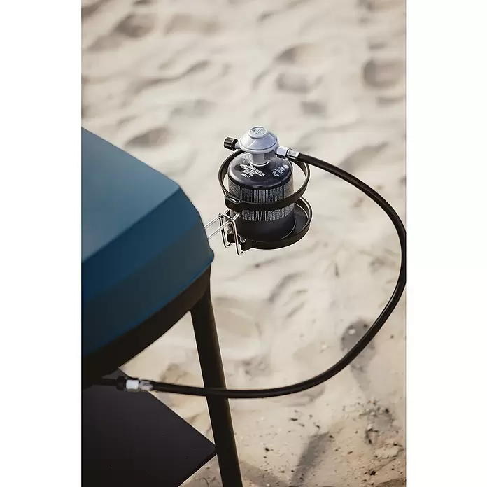 Halterung für Gaskartuschen - Passend für: Enders Urban II- und Explorer II-Serie - Schwarze Gaskartusche mit silbernem Regler und Schlauch, an einem dunkelblauen Grill auf Sand angeschlossen.