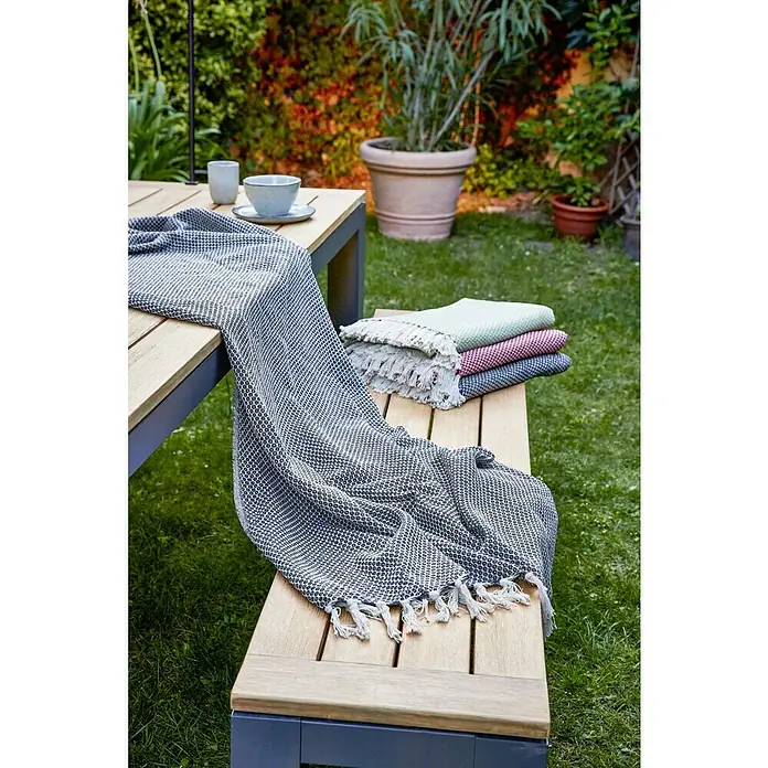 Sunfun Gartenbank Lynn - B x T x H: 195 x 40 x 45 cm, Naturbraun/Anthrazit - Gemütliche Gartenszene mit Tisch, Bank und Textilien: Eine dunkelgraue Tagesdecke mit Fransen liegt auf der Bank, daneben liegen weitere farbige Decken.