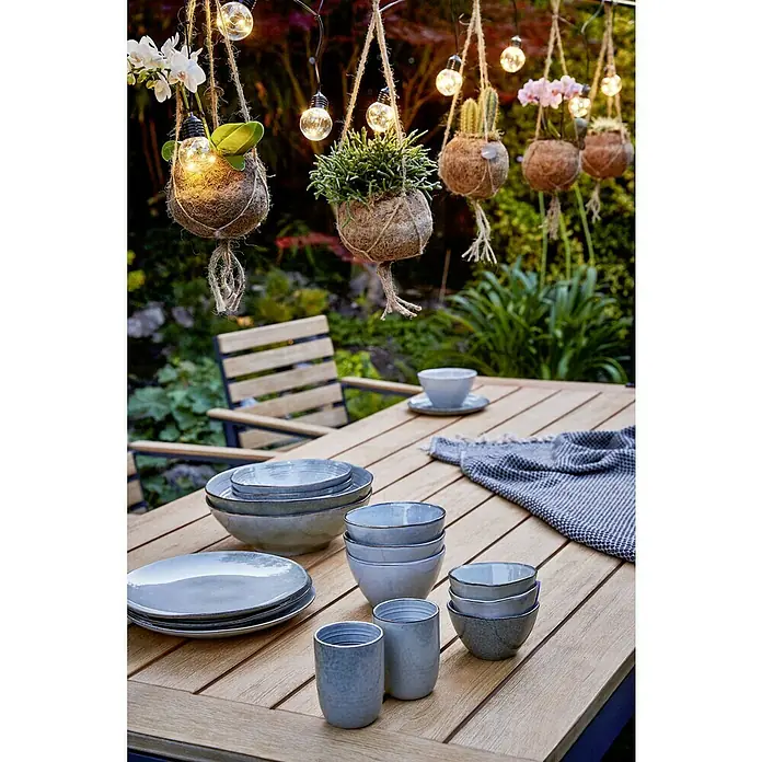 Sunfun Carina Silla de jardín - Acacia, Apilable, Natural, An x Pr x Al: 57,5 x 61 x 86 cm - Una acogedora mesa de jardín con vajilla, incluyendo platos, cuencos y tazas, con plantas colgantes y luces al fondo.
