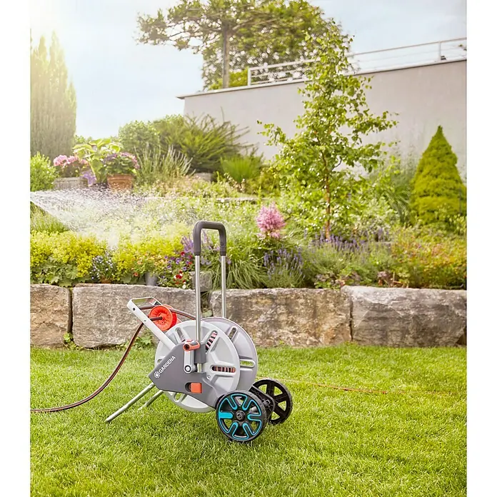 Gardena Schlauchwagen-Set CleverRoll M - Kunststoff/Metall, Schlauchlänge: 25 m - Eine Gardena Schlauchbox auf Rädern steht auf einem Rasen vor einem bunten Garten.