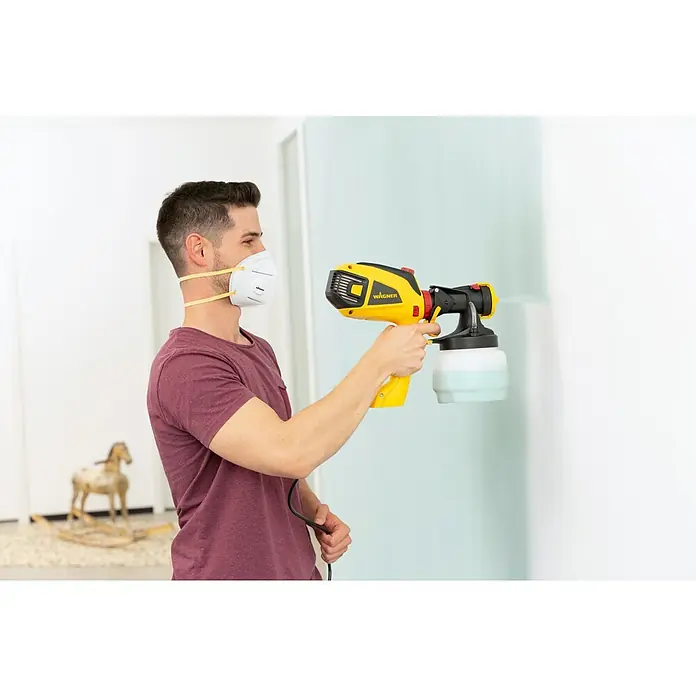 Wagner Farbsprühsystem W 590 Flexio - 630 W, Förderleistung: 0,5 l/min - Ein Mann mit Atemschutzmaske benutzt eine Wagner Farbspritzpistole, um eine Wand zu streichen. Das gelb-schwarze Sprühgerät sorgt für einen gleichmäßigen Farbauftrag.