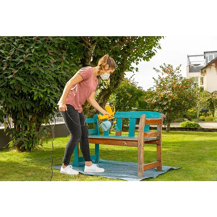 Wagner Farbsprühsystem W 590 Flexio - 630 W, Förderleistung: 0,5 l/min - Eine Frau lackiert eine Gartenbank im Freien mit einer gelben Farbspritzpistole und trägt dabei eine Maske.