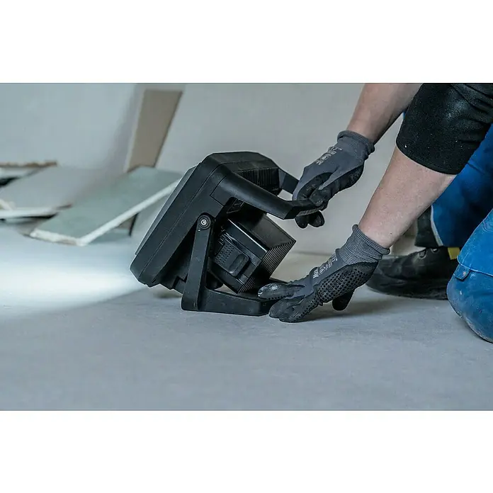 Brennenstuhl LED-Akkustrahler Professional Multi Battery - 3.150 lm, 6.500 K, 16,9 x 28,5 x 22,7 cm, IP65 - Nahaufnahme einer schwarzen Arbeitsleuchte, die auf dem Boden steht und von einer Person mit grauen Handschuhen eingestellt wird.