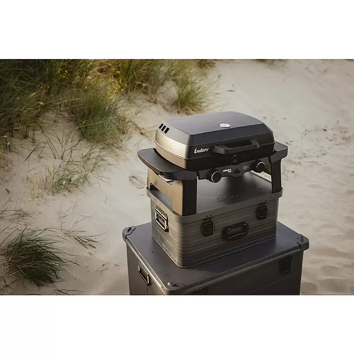 Enders Gasgrill Urban II - Grillfläche (B x T): 49 x 32 cm, 4,4 kW, Anzahl Brenner: 2 Stk. - Schwarzer tragbarer Gasgrill "Enders URBAN II PRO" auf zwei dunkelgrauen Aufbewahrungsboxen an einem Sandstrand mit Gras.