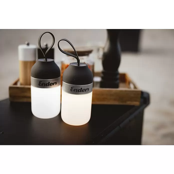 Enders Campinglampe Luvo - 100 lm - Zwei tragbare Enders Laternen, oben schwarz, mit leuchtendem weißem Korpus und perforierten Metallbändern, auf einem Tisch.