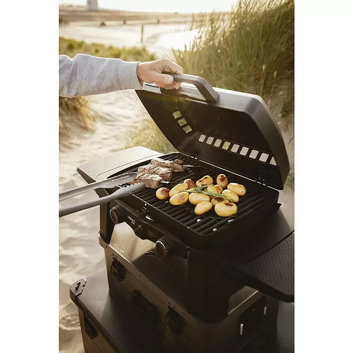 Enders Gasgrill Urban II - Grillfläche (B x T): 49 x 32 cm, 4,4 kW, Anzahl Brenner: 2 Stk. - Schwarzer tragbarer Grill, Enders Urban II Pro, gart Fleisch und Kartoffeln am Sandstrand, von einer Person gehalten. Auf dunklem Koffer.