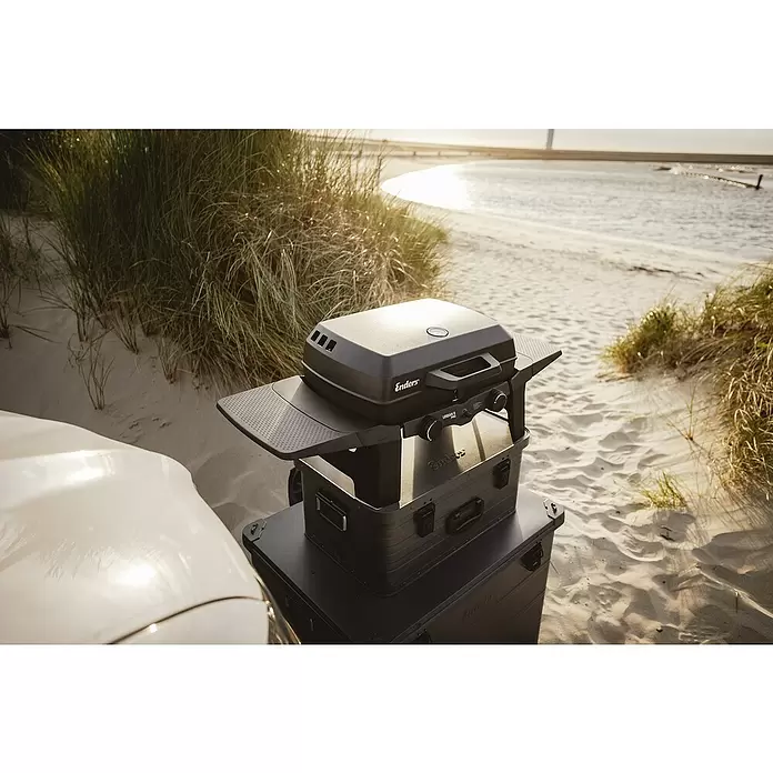 Enders Gasgrill Urban II - Grillfläche (B x T): 49 x 32 cm, 4,4 kW, Anzahl Brenner: 2 Stk. - Schwarzer Enders Urban II Pro Gasgrill auf gestapelten Koffern mit ausziehbaren Seitenablagen am Sandstrand.