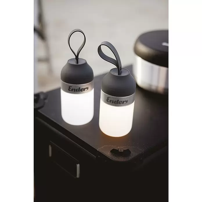 Enders Campinglampe Luvo - 100 lm - Zwei Enders Laternen mit schwarzen Oberteilen und silbernen Lochbändern, eine mit weißem, eine mit warmem Licht, auf dunklem Untergrund.