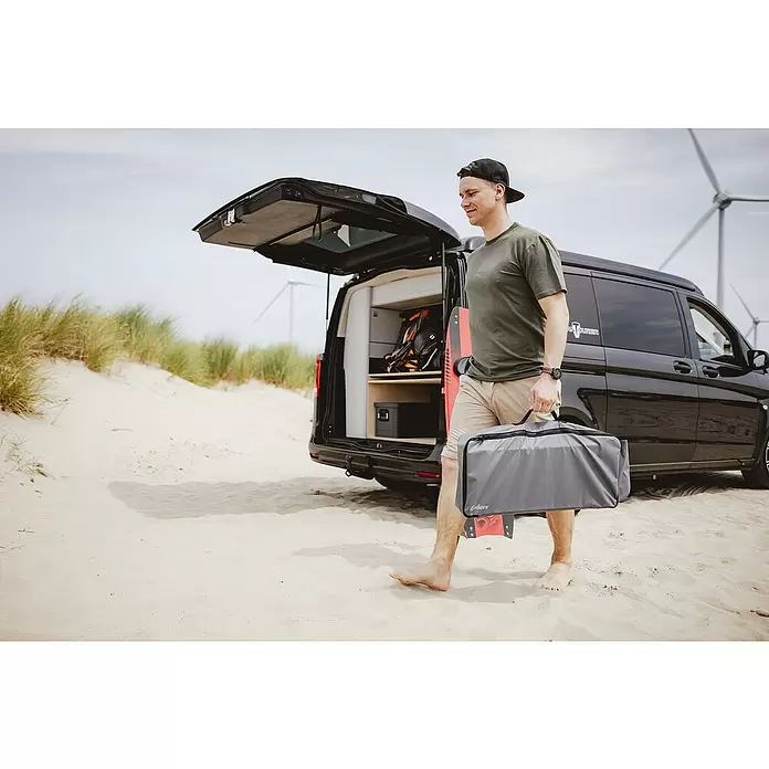 Transporttasche - Passend für: Enders Gaskocher Bondy 2, PVC - Mann trägt eine graue Reisetasche mit schwarzem Reißverschluss an einem Sandstrand neben einem Campervan.