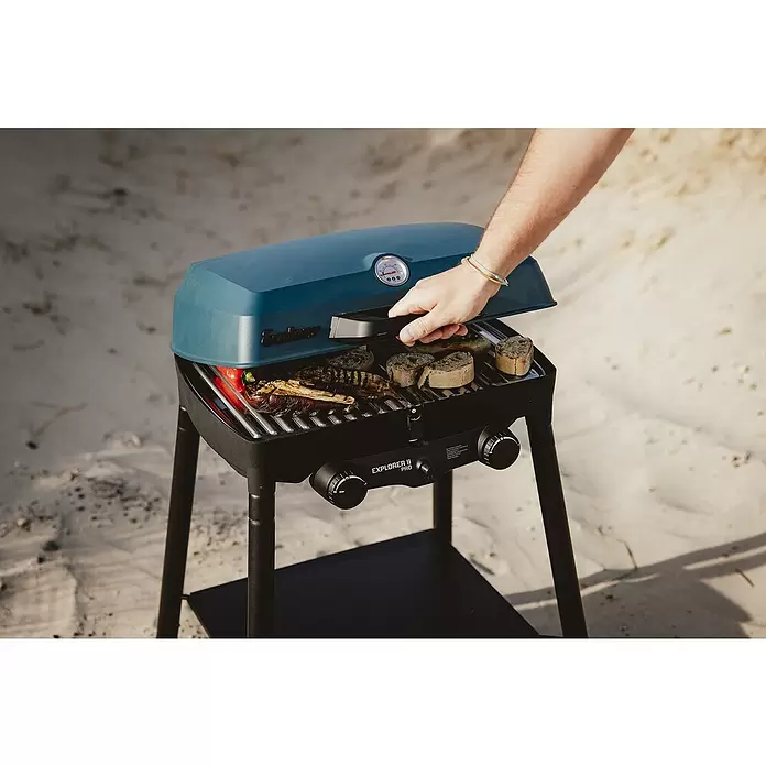 Enders Gasgrill Explorer II Pro - Grillfläche (B x T): 49 x 32 cm, 4,4 kW, Anzahl Brenner: 2 Stk., 50 mbar - Ein teal Enders Explorer II Pro Gasgrill kocht Gemüse und Brot, eine Hand hebt den Deckel auf sandigem Untergrund.