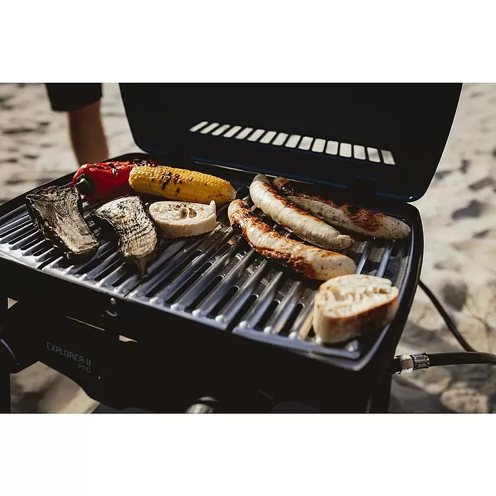 Enders Gasgrill Explorer II Pro Caravan - Grillfläche (B x T): 49 x 32 cm, 4,4 kW, Anzahl Brenner: 2 Stk., 30 mbar - Schwarzer tragbarer Grill Explorer II Pro, der draußen auf Sand Gemüse, Würstchen und Brot zubereitet.