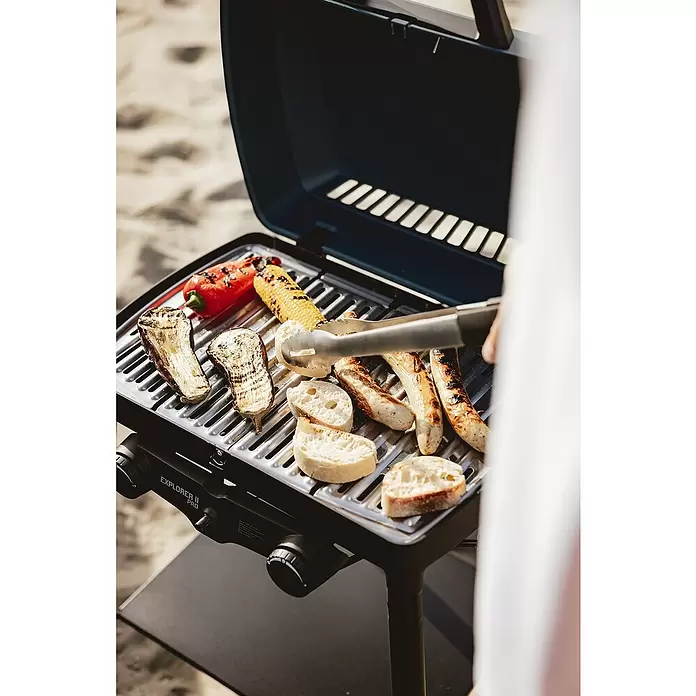 Enders Gasgrill Explorer II Pro - Grillfläche (B x T): 49 x 32 cm, 4,4 kW, Anzahl Brenner: 2 Stk., 50 mbar - Eine Person grillt auf einem schwarzen tragbaren Gasgrill Auberginen, Paprika, Mais, Würstchen und Brot auf sandigem Untergrund.